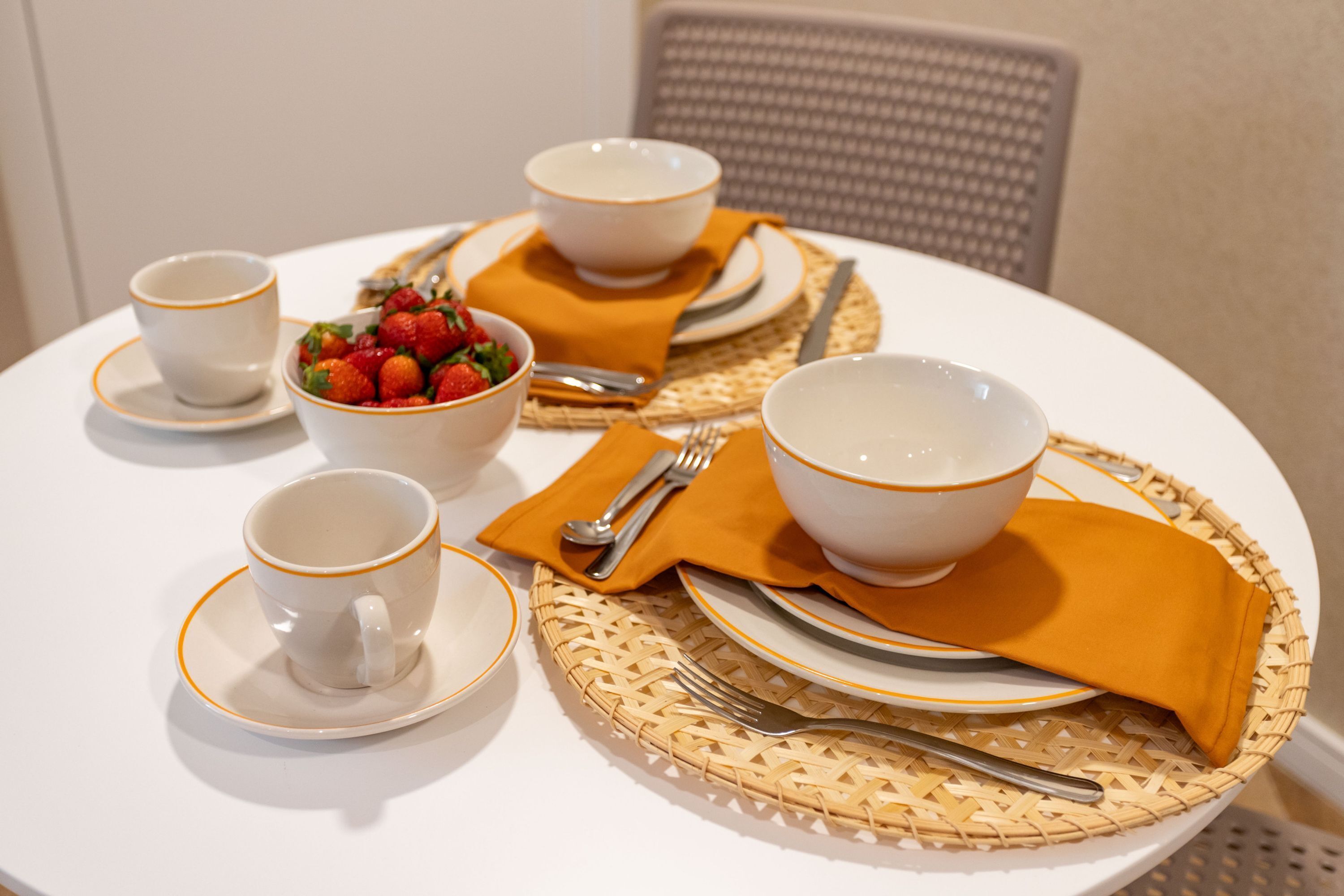 A round dining table with white plates, bowls, and cups, accompanied by a woven placemat and a mustard-colored napkin.