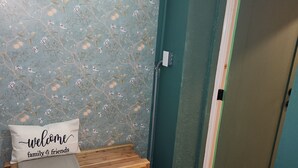 Une pièce avec un papier peint à motifs floraux, un banc agrémenté d’un coussin sur lequel est écrit « Bienvenue à la famille et aux amis », et une porte dotée d’un cadre vert.