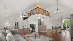 Amplia sala de estar con un sofá grande de color gris, una mesa de comedor con sillas, una zona de cocina y una escalera que lleva a un nivel superior.