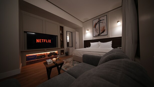 A modern hotel room with a large flat-screen TV displaying 'NETFLIX', a neatly made bed, a sofa, and a small table with a remote control. - Nma a Residences 2 (Riyadh)