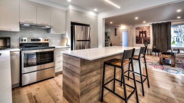 A modern kitchen with stainless steel appliances, a wooden island with bar stools, and a view of the living area through an open door. - Lux. 4BR House with Pool near DT (Brossard)