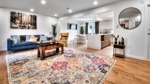 A spacious living room with a blue sofa, a coffee table, and a colorful rug. There is a kitchen area with a dining table and chairs visible in the background. - Lux. 4BR House with Pool near DT (Brossard)