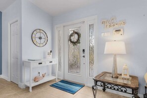 Un couloir avec une console blanche, une horloge, une porte dotĂ©e dâun panneau en verre dĂ©poli, un panneau dâaccueil, une lampe et une petite table avec une lampe.