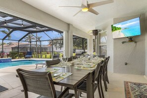Un espace extérieur abrité, aménagé pour les repas, avec une piscine, un ventilateur de plafond et un téléviseur fixé au mur.