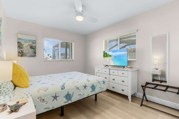 Un dormitorio con cama, cómoda, televisor, ventana con vista a la playa y ventilador de techo.