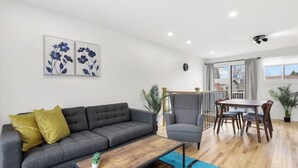 A modern living room with a grey sofa, yellow cushions, a wooden coffee table, and a grey armchair. There is a dining area with a table and chairs, and a balcony with a railing. - Elegant 4 bedroom modern house (Brossard)