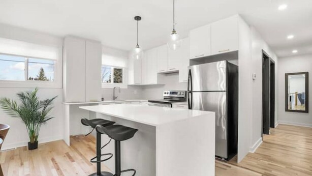 A modern kitchen with white cabinetry, a stainless steel refrigerator, a large island with bar stools, and pendant lighting. - Elegant 4 bedroom modern house (Brossard)