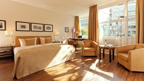 A hotel room with a large bed, two armchairs, a desk, and a view of buildings through large windows. - Abion Villa Suites (Berlin)