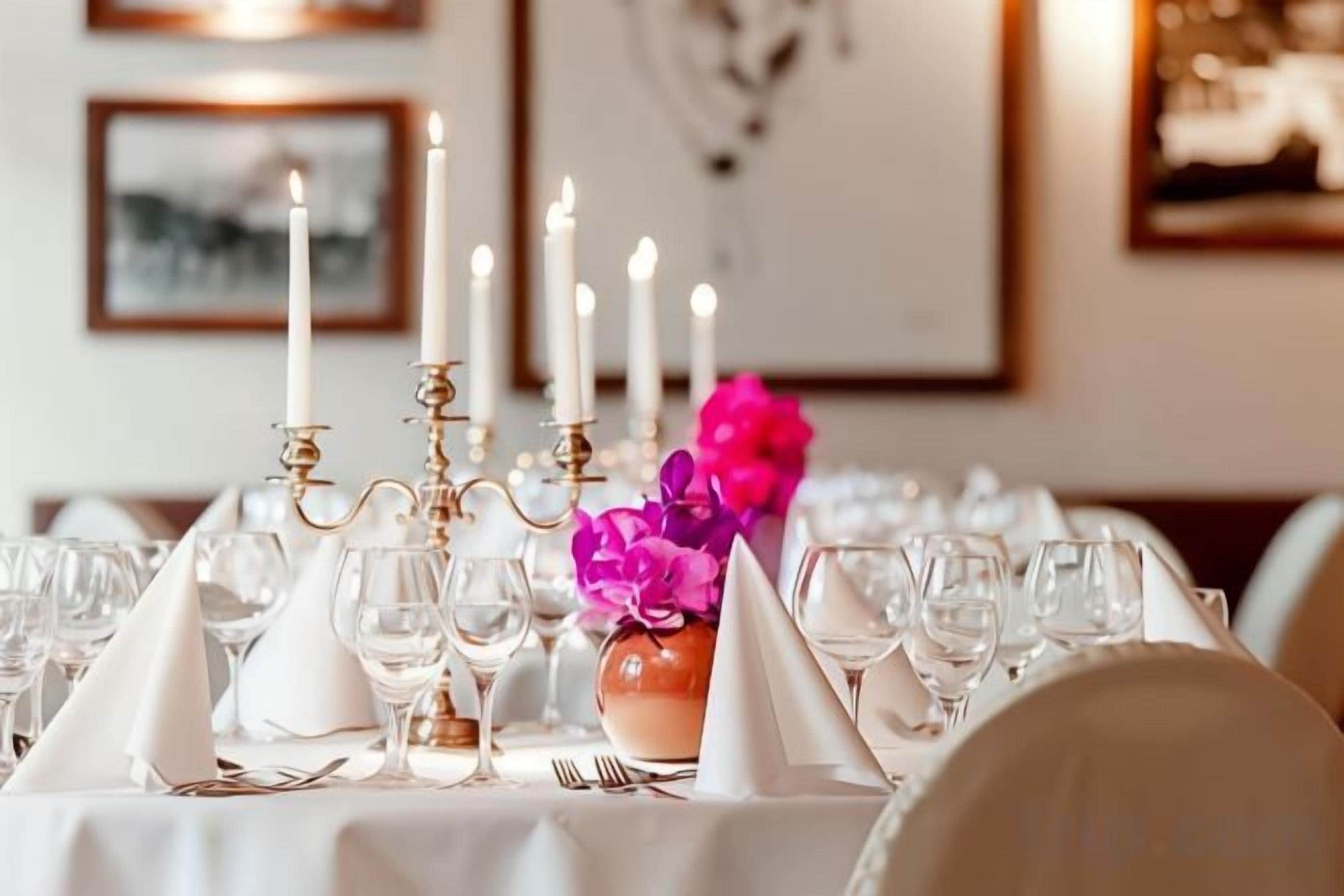 Ein gedeckter Tisch mit mehreren Weingläsern, einer Vase mit rosa Blumen und brennenden Kerzen.