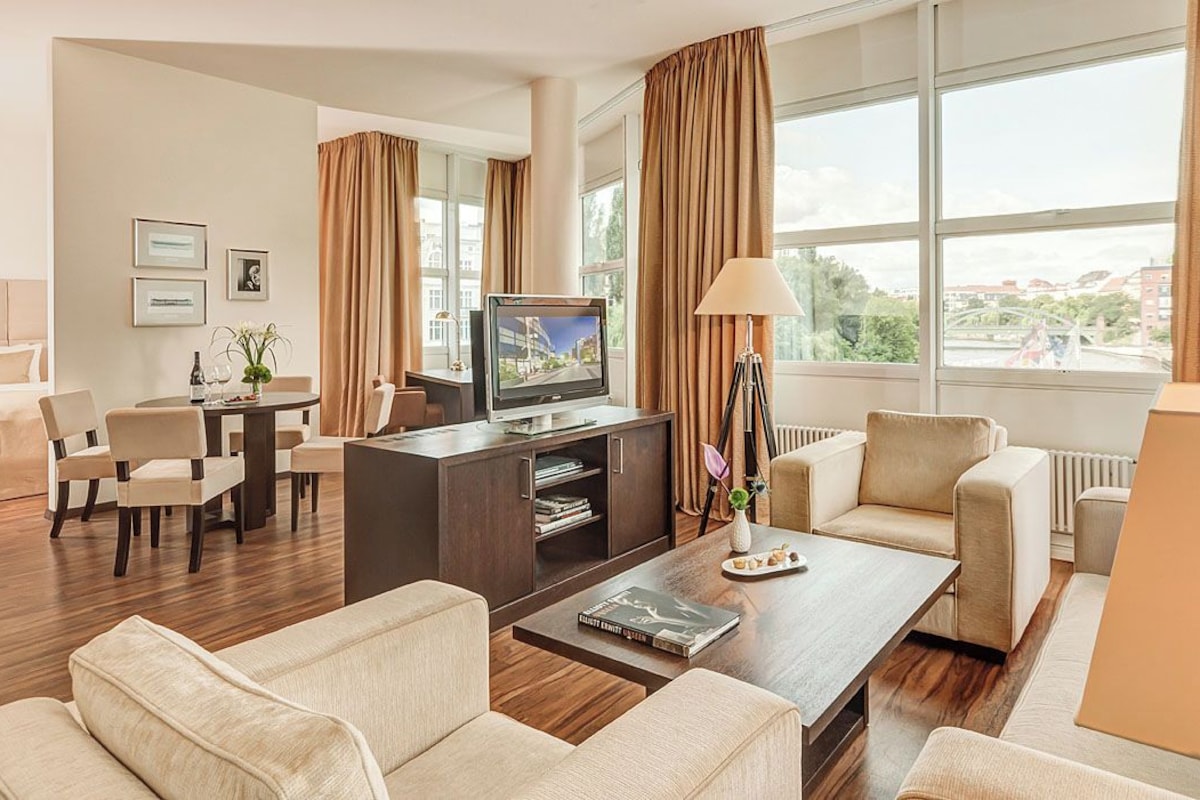 A hotel room with a large window, a television, a dining area, and a sofa.