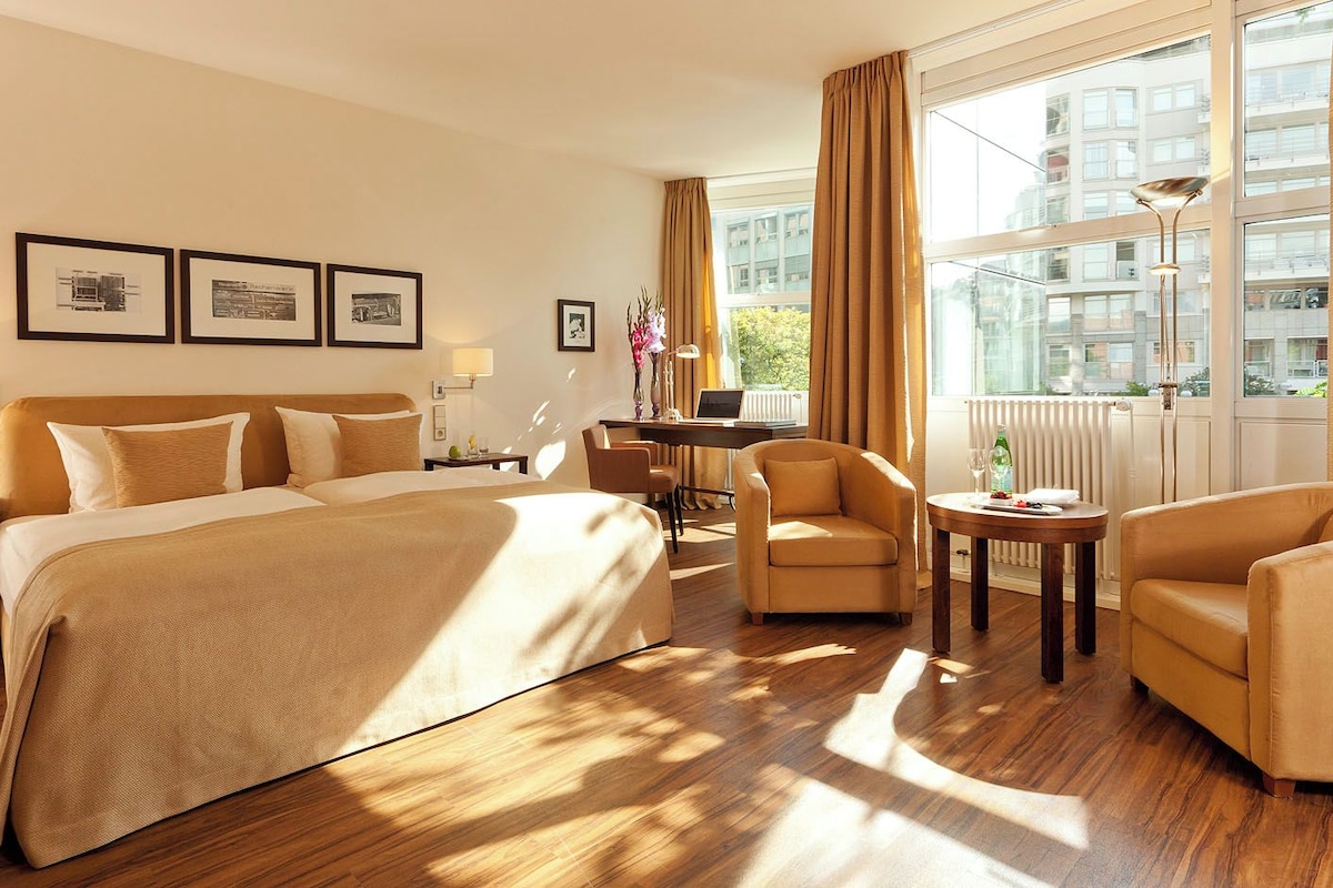 A hotel room with a large bed, two armchairs, a small table, and a desk with a computer. There are framed pictures on the wall, a window with curtains, and a radiator.