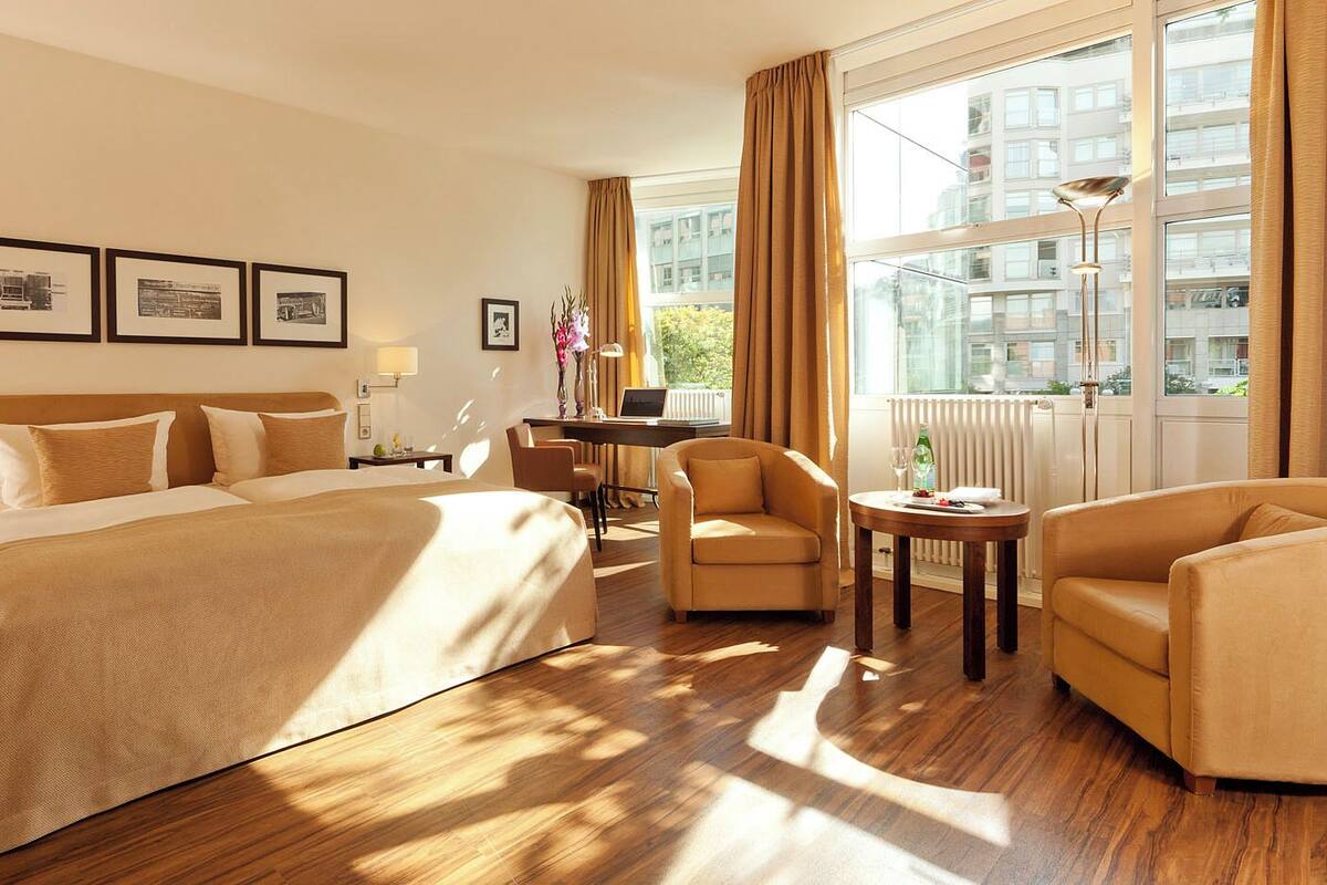 A hotel room with a large bed, two armchairs, a small table, and a desk with a computer. There are framed pictures on the wall, a window with curtains, and a radiator.