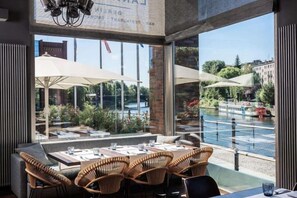 An outdoor dining area with wicker chairs and tables, large umbrellas, and a view of a river and buildings. - Abion Villa Suites (Berlin)