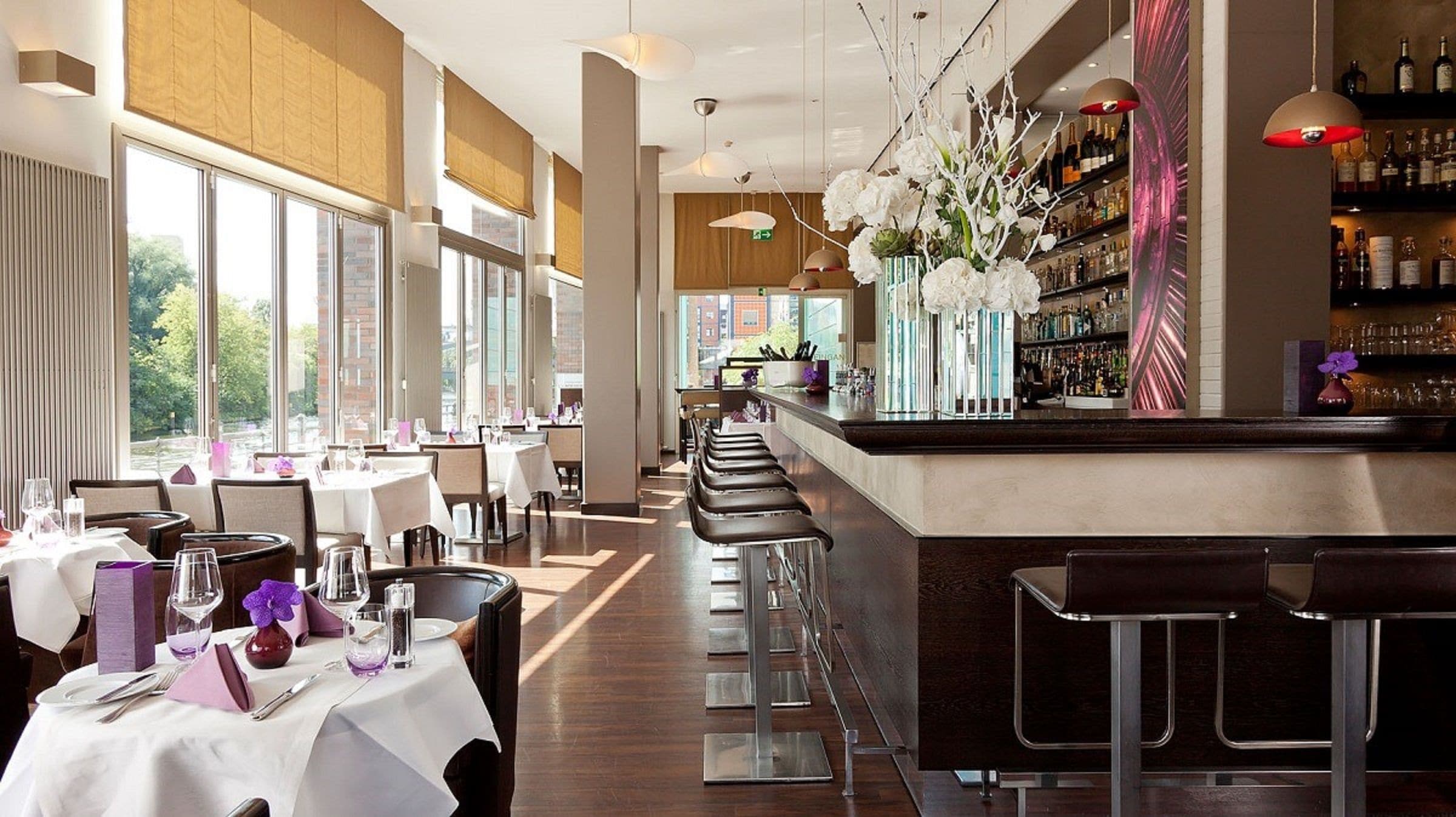 A modern restaurant with a long bar, white tablecloths, and a view of the city.