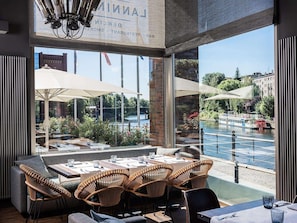 An outdoor dining area with wicker chairs and tables, overlooking a river with boats and greenery. - Abion Villa Suites (Berlin)