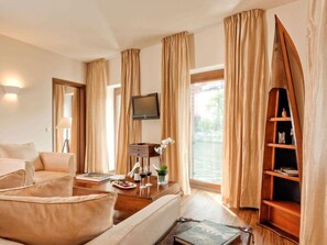 A spacious living room with a sofa, a coffee table, and a television. There is a window with curtains, a bookshelf, and a view of a body of water. - Abion Villa Suites (Berlin)