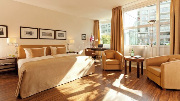 A hotel room with a large bed, two armchairs, a small table, and a desk with a computer. There are framed pictures on the wall, a window with curtains, and a radiator. - Abion Villa Suites (Berlin)