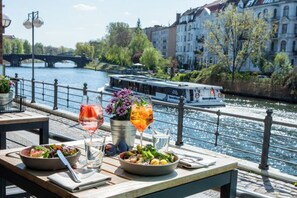 A riverside dining table with food and drinks, a boat on the water, and buildings in the background. - Abion Villa Suites (Berlin)