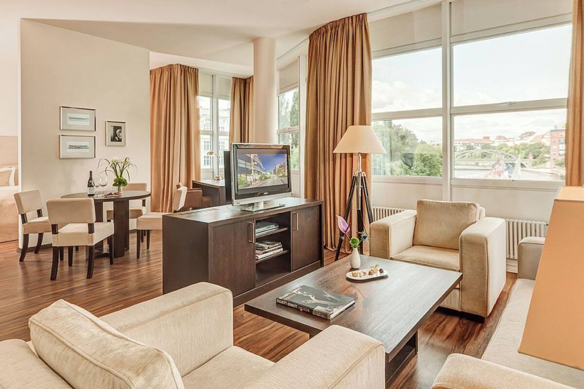 A hotel room with a large window, a television, a dining area, and a sofa.