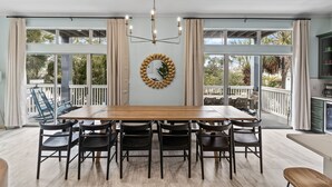 A dining area with a long wooden table and six chairs, a chandelier, and a large window with curtains.