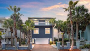 A two-story residential building with a central garage, surrounded by palm trees and a brick driveway.