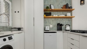 Ett modernt kök med vita skåp, en inbyggd tvättmaskin, ett kompakt kylskåp och hyllor i trä med köksredskap.