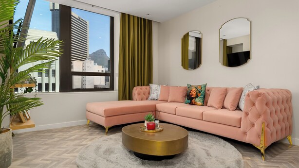 Een moderne woonkamer met een roze bank, een ronde gouden salontafel en een groot raam met uitzicht op gebouwen.