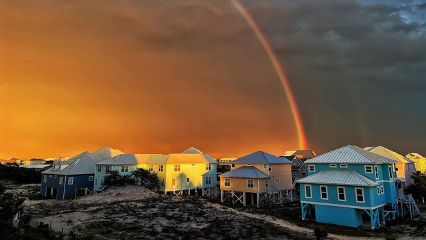 Una propiedad frente a la playa con casas elevadas sobre pilotes, una playa de arena y un arcoíris en el cielo.