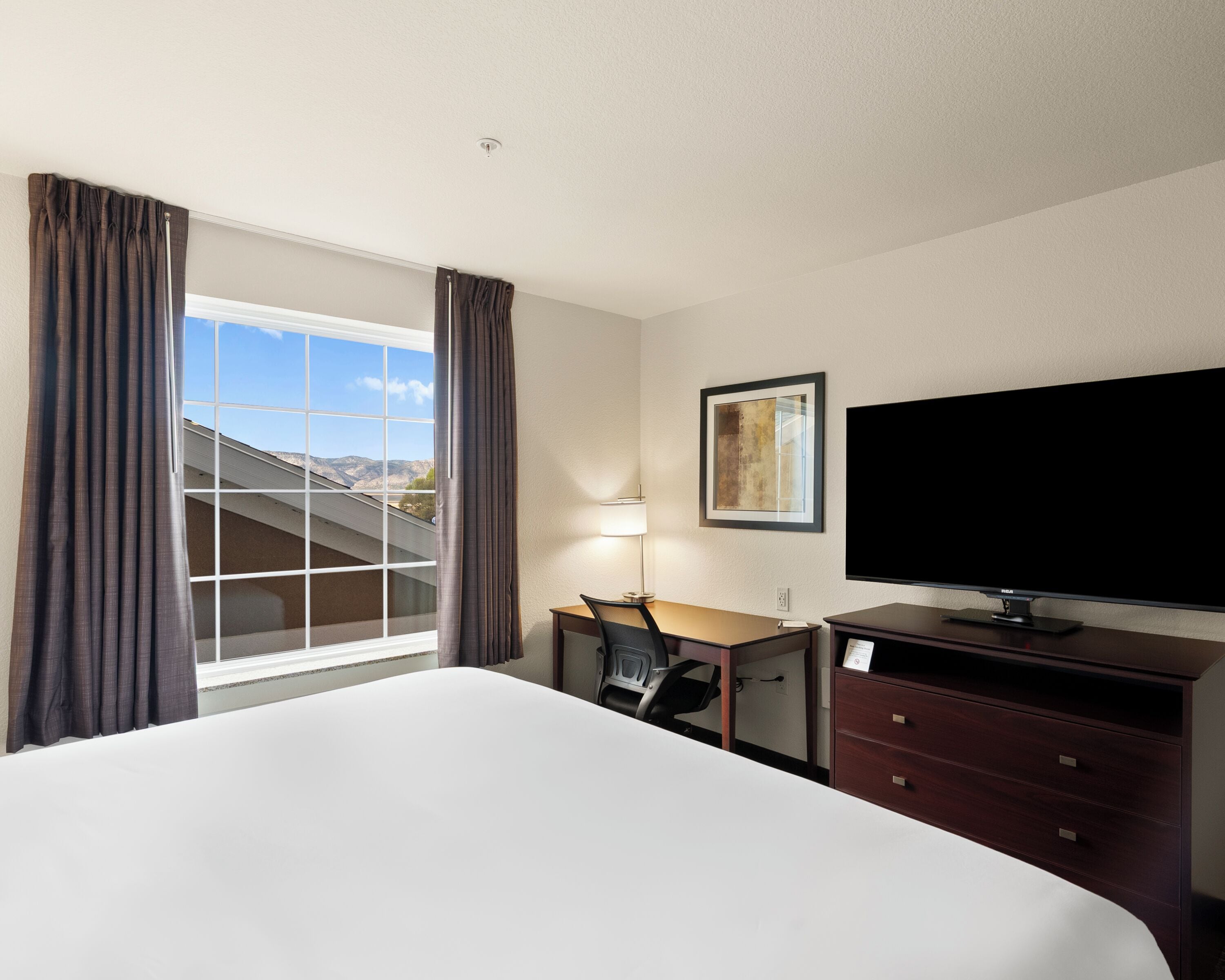 Quarto de hotel com uma cama grande, uma escrivaninha, televisão e janela com cortinas.