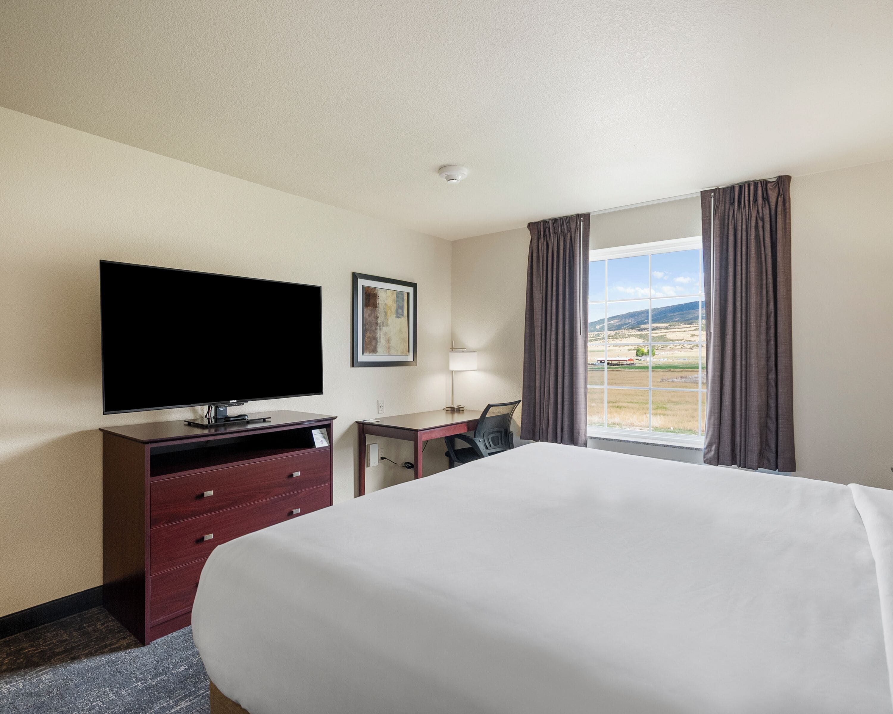 A hotel room with a large bed, a flat-screen TV, a desk, and a window with curtains.