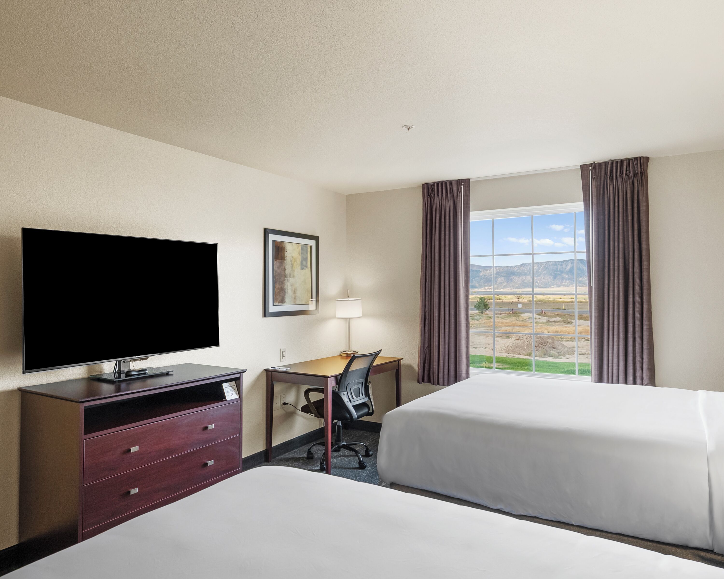 A hotel room with a flat-screen TV, a desk with a chair, two beds, a window with curtains, and a painting on the wall.