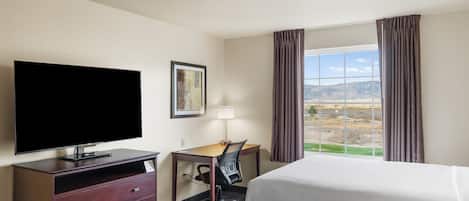 Quarto de hotel com televisão de tela plana, escrivaninha com cadeira, duas camas, janela com cortinas e um quadro na parede.