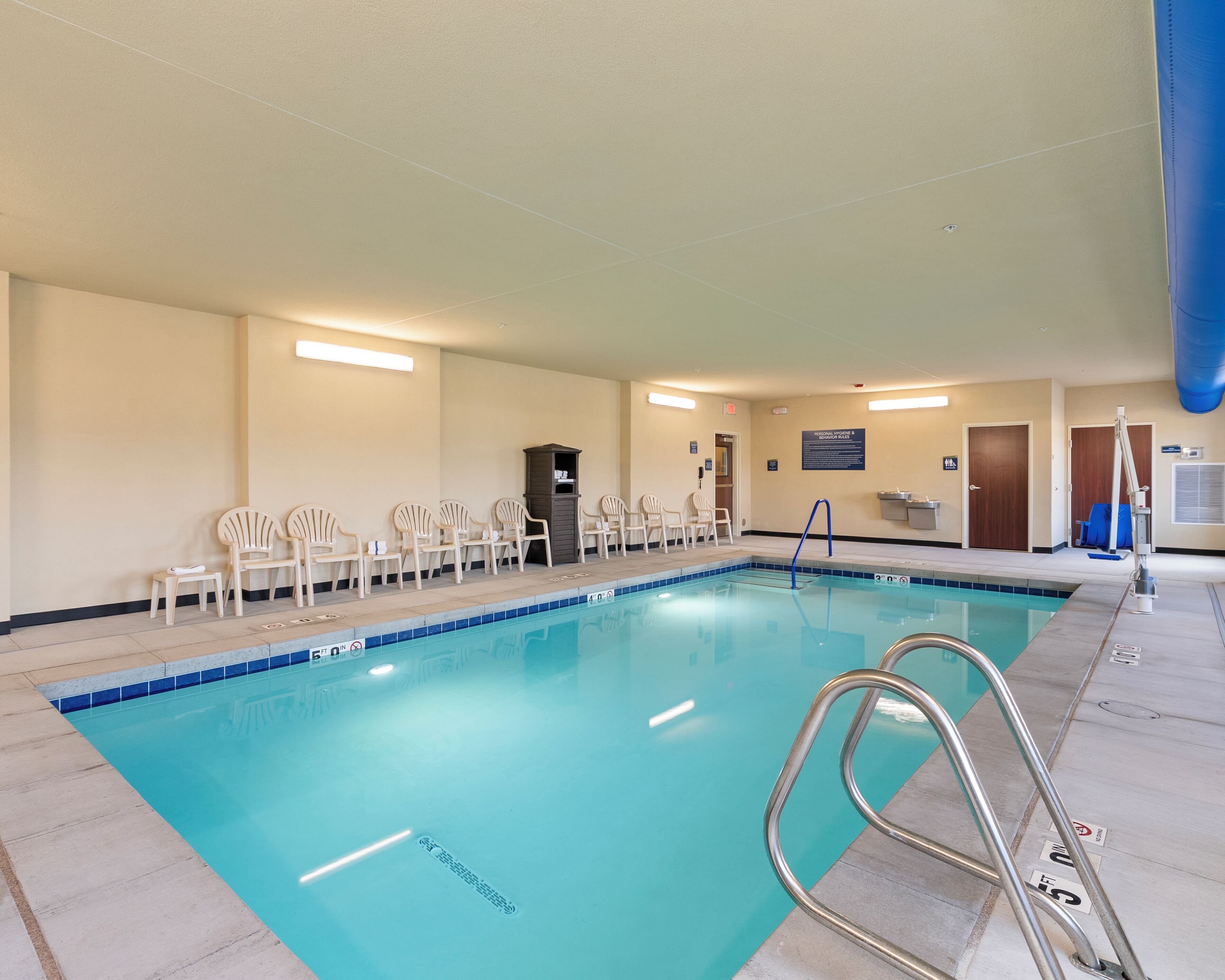 Una piscina interior con agua azul cristalina, rodeada de sillas blancas y una pequeña recepción.