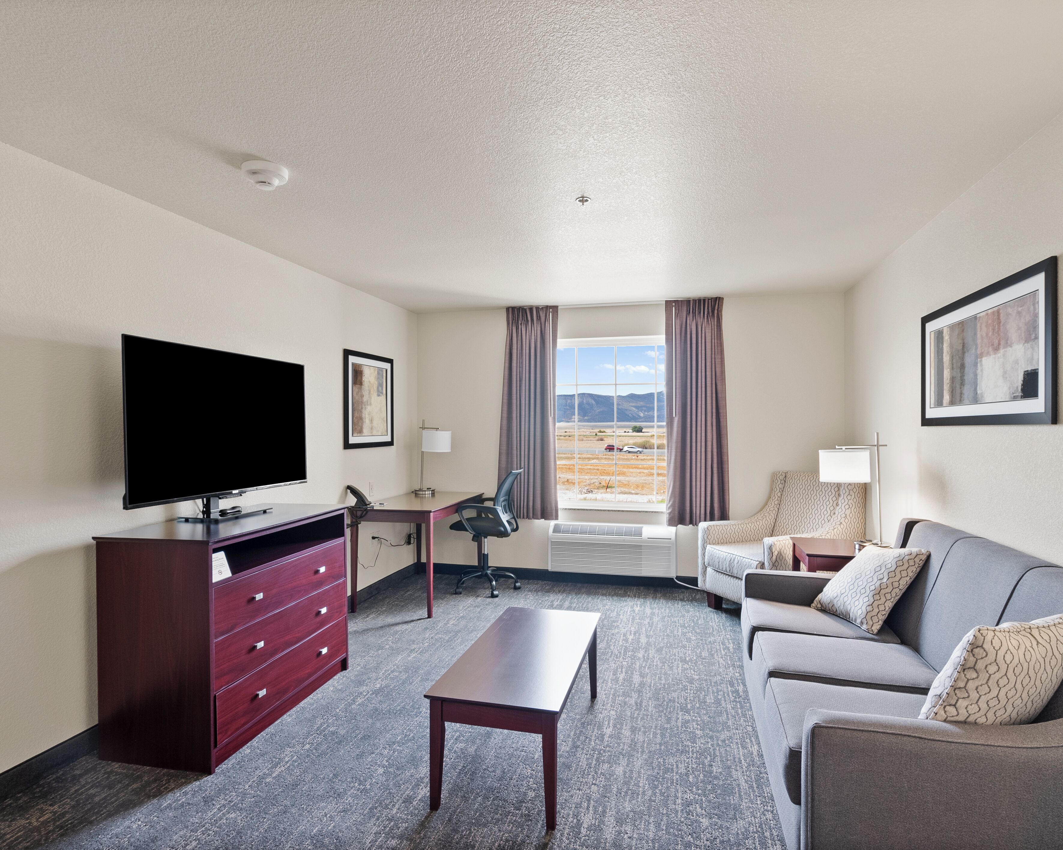 Une chambre d’hôtel équipée d’une télévision à écran plat, d’un bureau avec une chaise, d’un canapé, d’une commode et d’une grande fenêtre offrant une vue sur les montagnes.