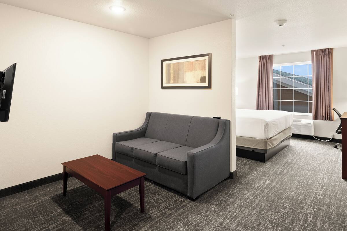 A hotel room with a gray sofa, a wooden table, a bed, and a window with curtains.