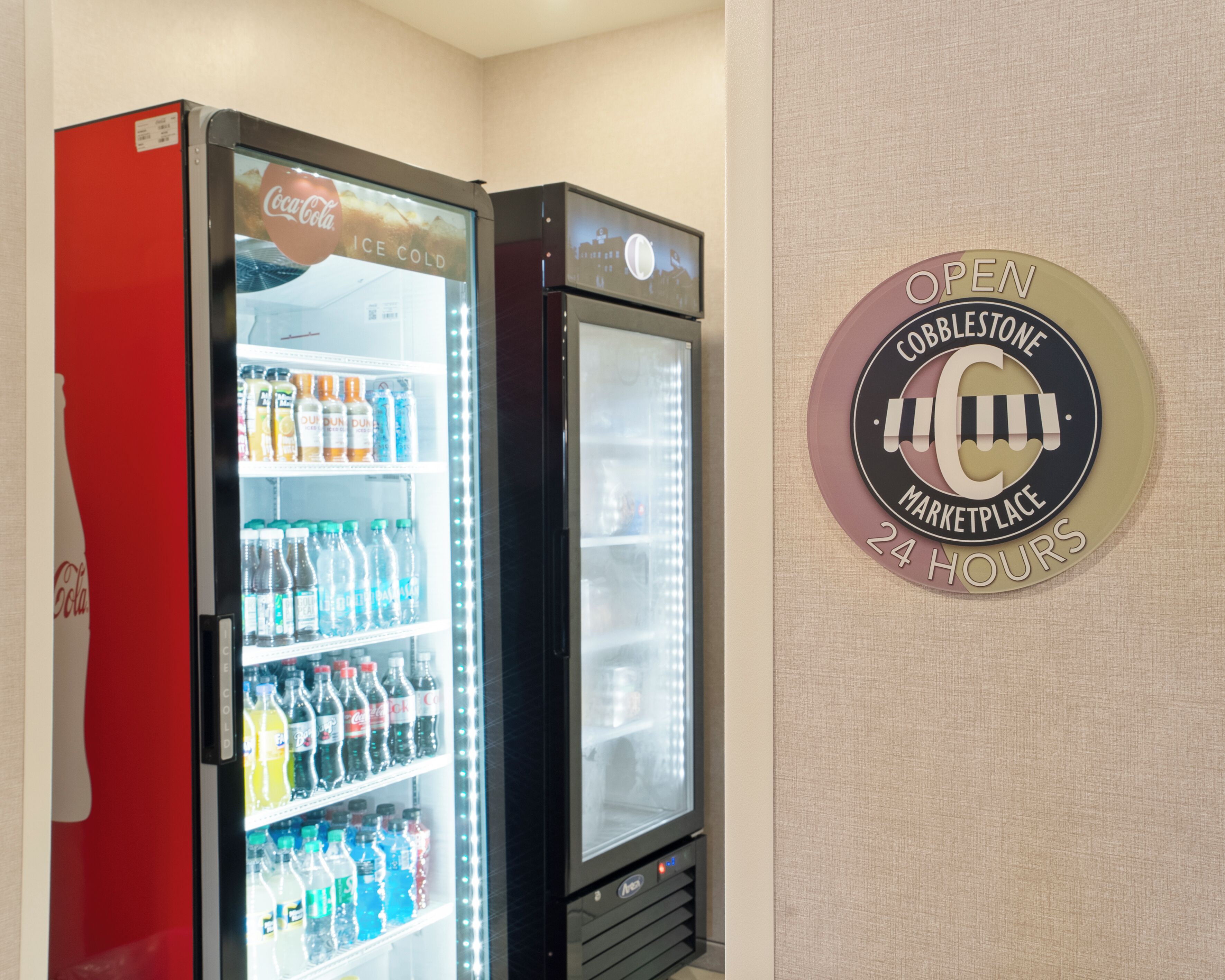 Two beverage vending machines with a sign indicating the location is a Cobblestone Marketplace open 24 hours.
