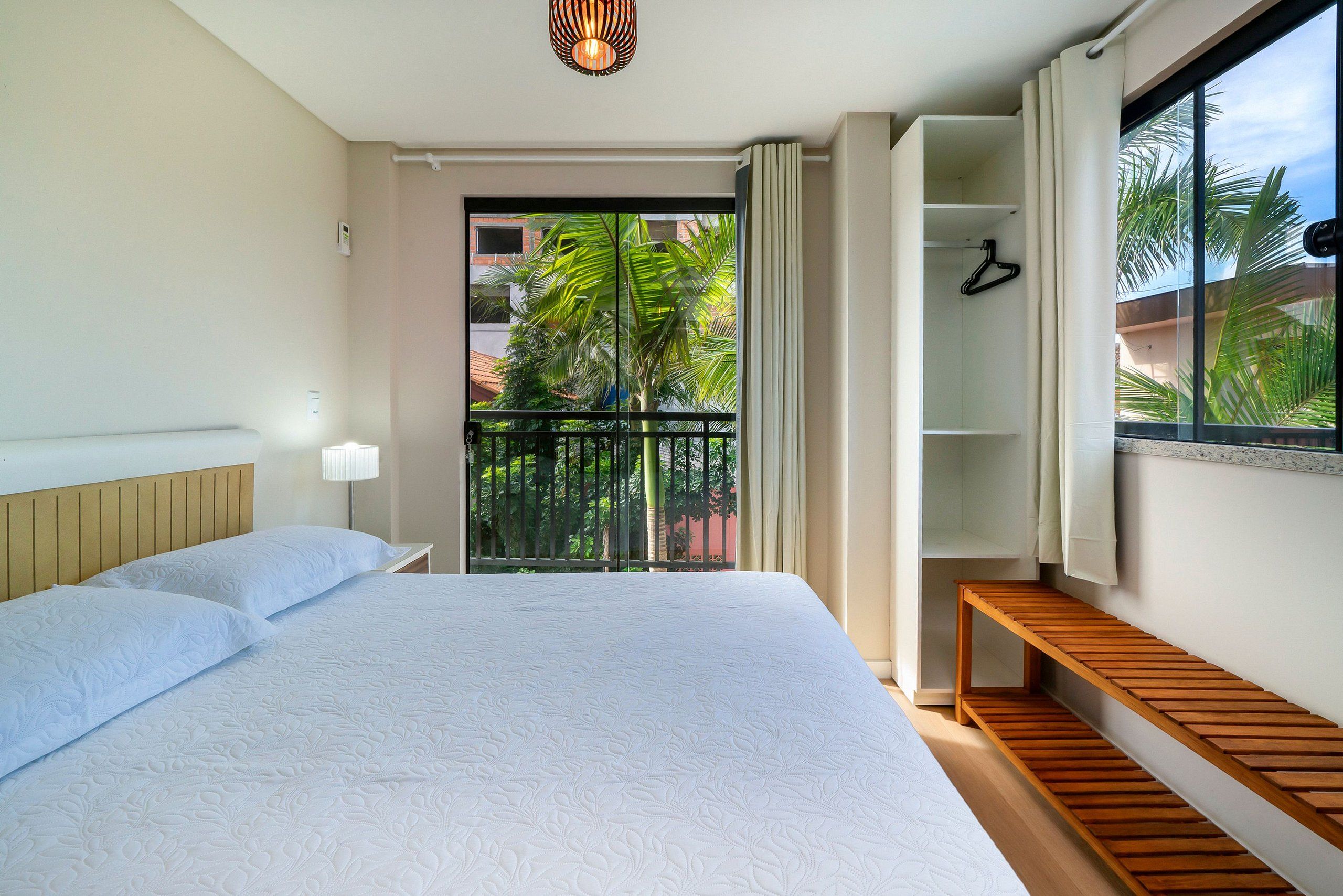 Habitación de hotel con una cama grande, un banco de madera, un armario y un balcón con vistas a palmeras.