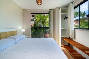 Une chambre d’hôtel avec un grand lit, un banc en bois, une armoire et un balcon donnant sur des palmiers.