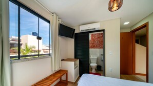 A hotel room with a bed, a wooden bench, a TV, an air conditioning unit, and a view of buildings outside. - Suíte 8 Ala pôr sol Residencial Solavir (Bombinhas)