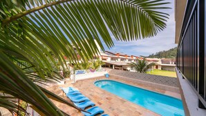 A swimming pool with blue lounge chairs, surrounded by palm trees and a view of buildings in the background. - Suíte 8 Ala pôr sol Residencial Solavir (Bombinhas)