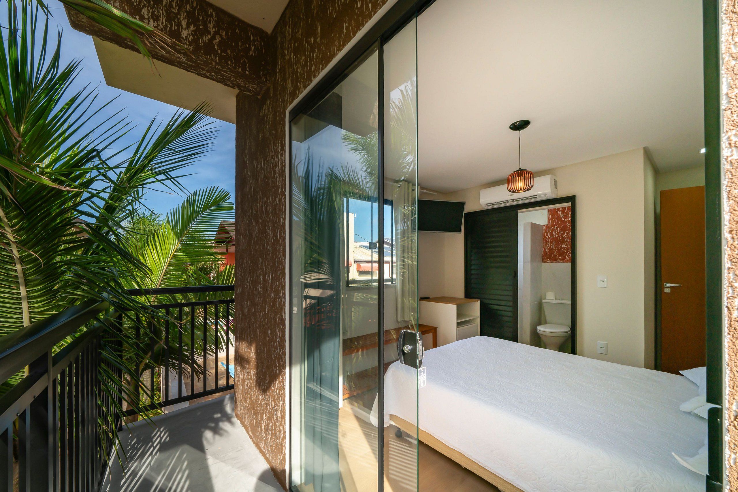 A modern bedroom with a large bed, a balcony with a glass door, and a view of palm trees.