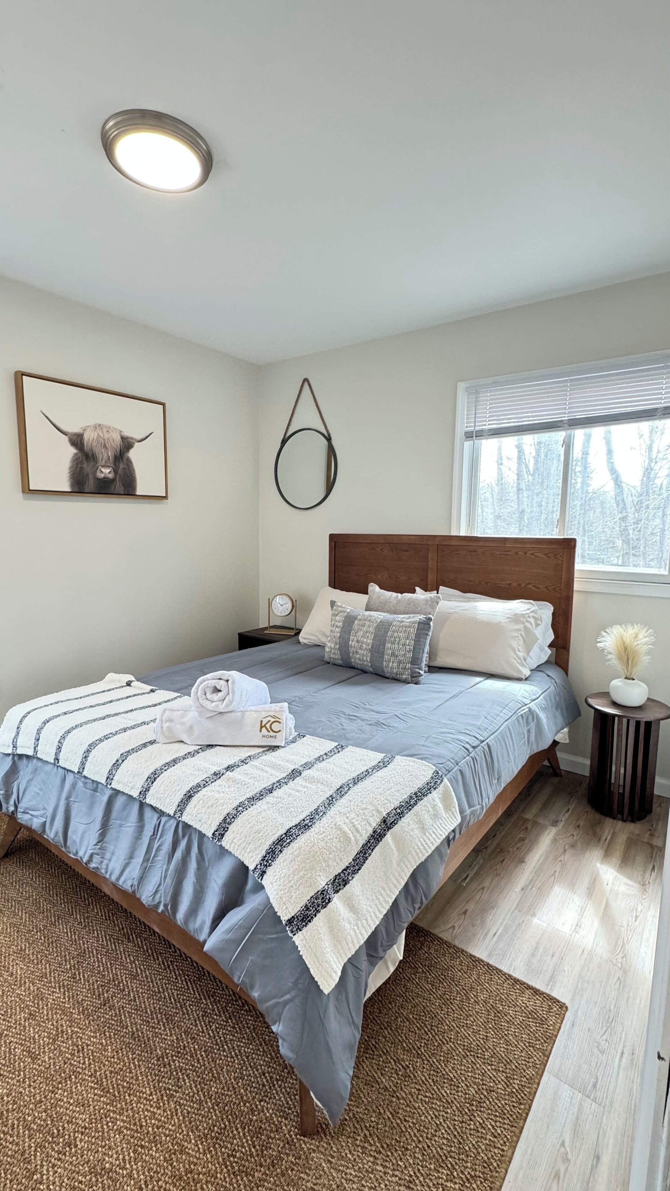 A bedroom with a bed, a bedside table, a clock, a framed picture, a window, and a hanging light fixture.