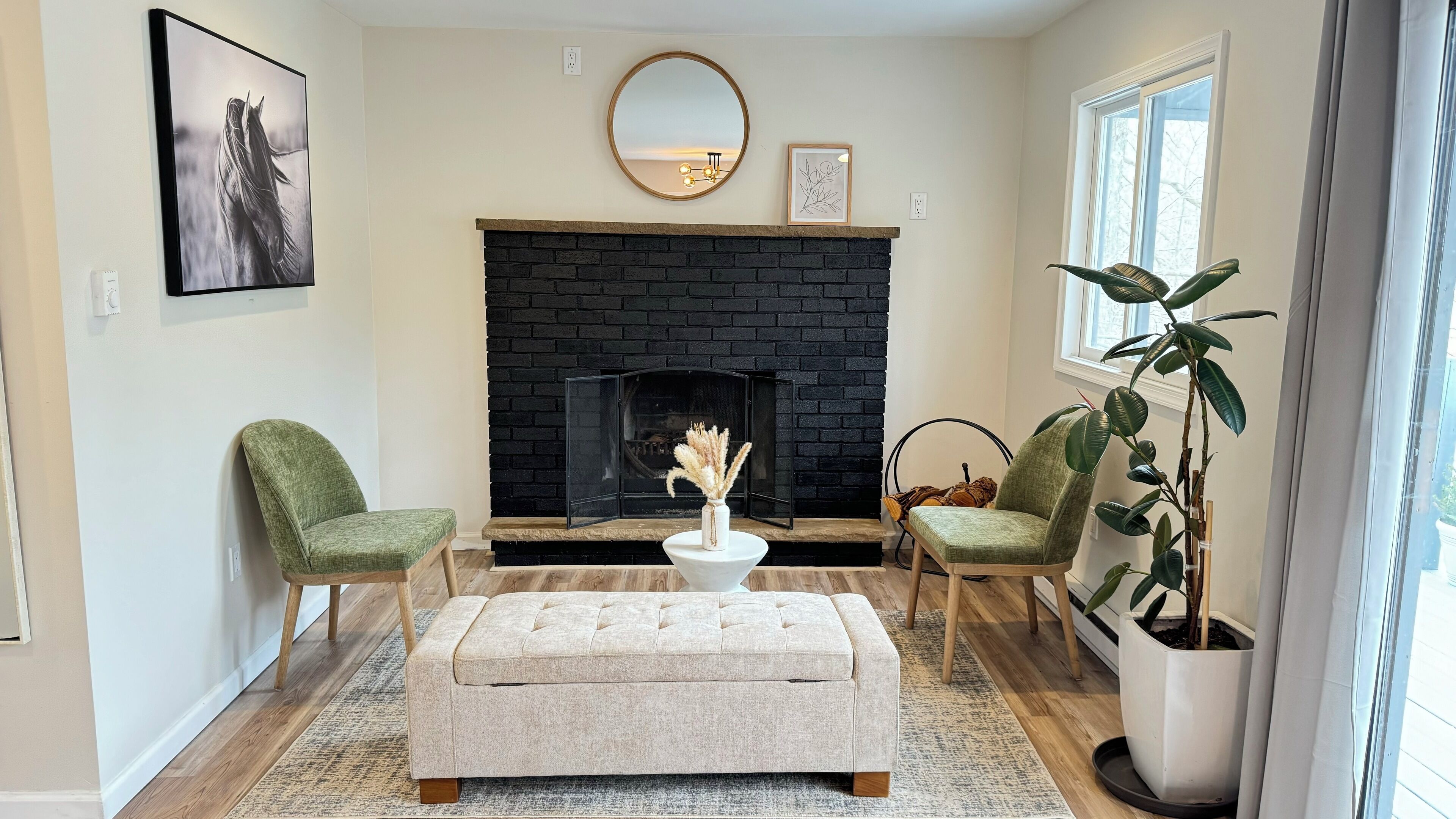 Un salon avec une cheminée, deux fauteuils verts, un pouf blanc et une grande plante en pot.