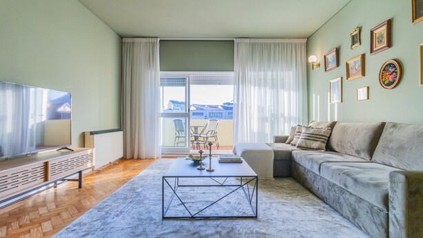 Ein modernes Wohnzimmer mit Sofa, Couchtisch, Fernseher und einem Balkon mit Gartenmöbeln.