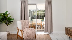 Habitación con una silla blanca, una planta grande en maceta y vistas a un área exterior con sillas y una mesa a través de una puerta de vidrio.