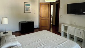 Un dormitorio con una cama grande, una mesita de noche con lámpara, un aparador, un televisor en la pared y una puerta que da a otra habitación.