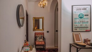 Un couloir orné d’un lustre, d’un miroir, d’un tableau encadré, d’une chaise avec un coussin et d’une table sur laquelle se trouve un plateau de collations.