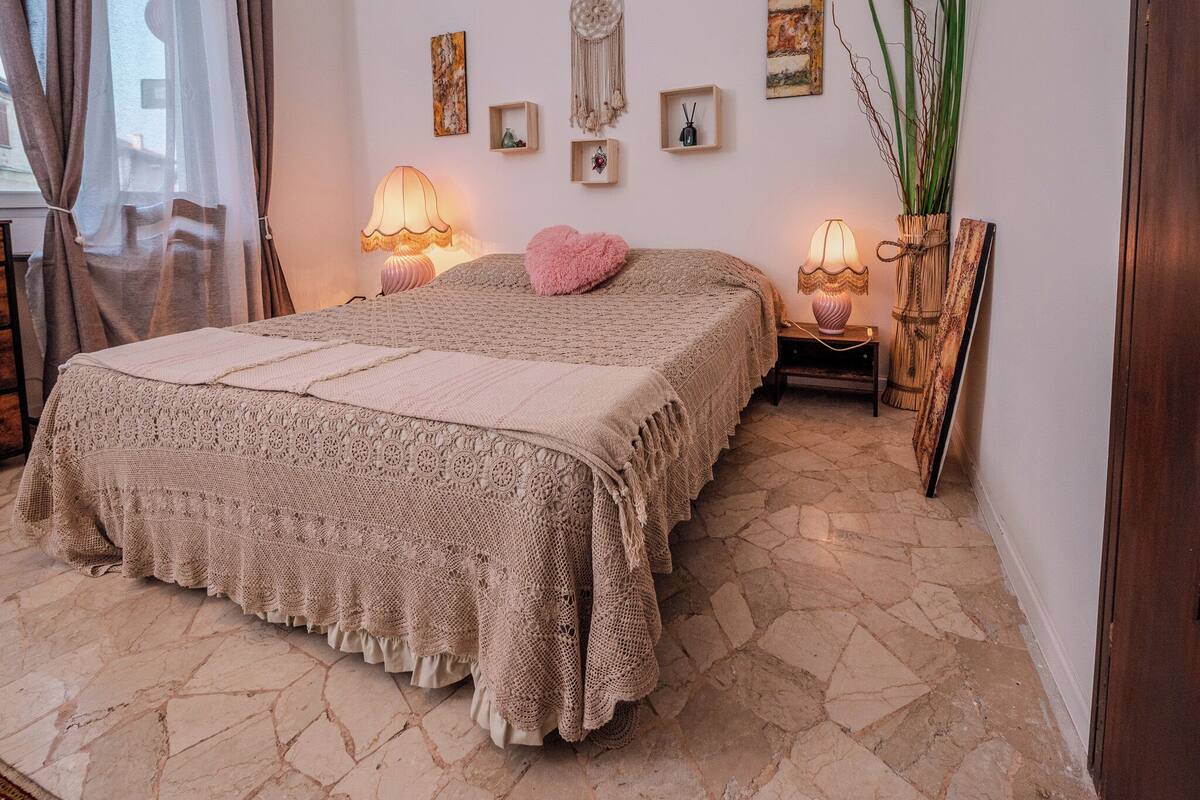 Ein gemütliches Schlafzimmer mit einem großen Bett, zwei Nachttischlampen, einem dekorativen Wandbehang aus Makramee und Steinböden.