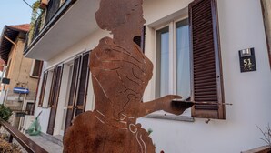 Une sculpture métallique représentant une femme sur un balcon, avec le numéro 51 d’une maison visible en arrière-plan.