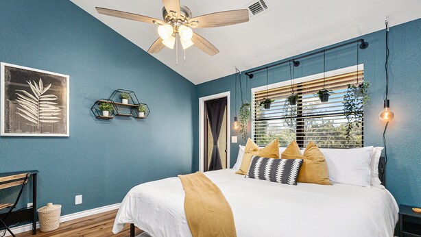 Ein Schlafzimmer mit einem Bett, einem Deckenventilator, einem Fenster mit Jalousien und einem an der Wand befestigten Regal mit Pflanzen.